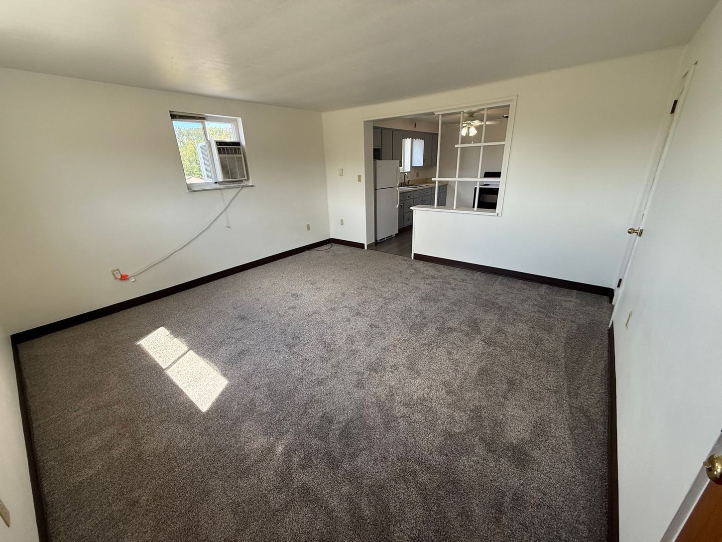 Empty living room with brown carpet and a view into the kitchen. White walls and a window air conditioner.