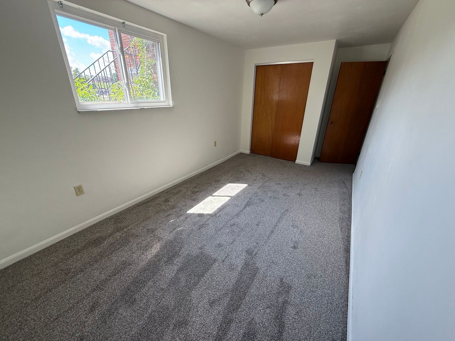 Empty room with gray carpet, white walls, two brown closet doors, and a window.