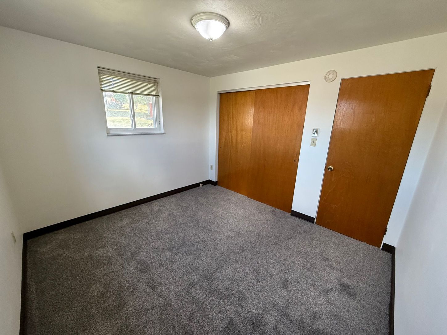 Empty room with gray carpet, two wooden doors, and a small window with a shade.