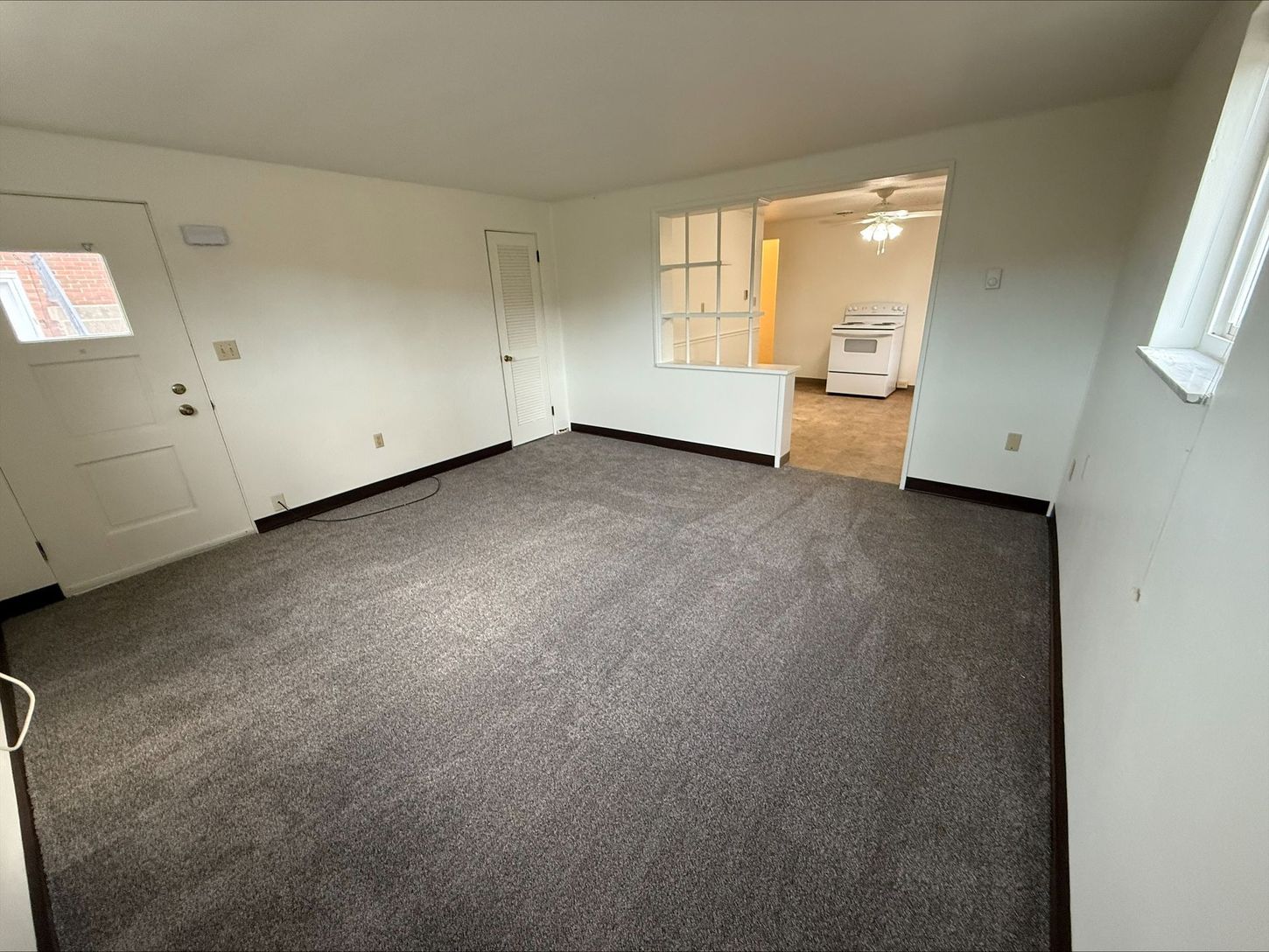 Empty living room with gray carpet, white walls, doorway to kitchen.