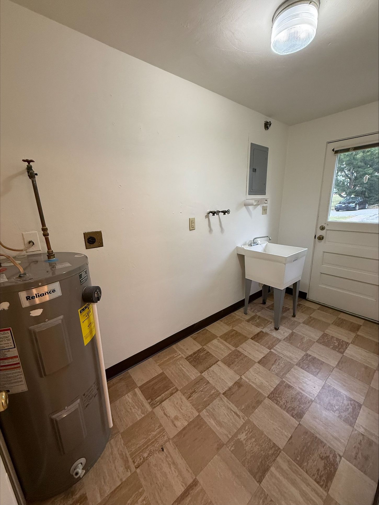 Laundry room: water heater, sink, electrical panel, open door, beige and brown tiled floor.