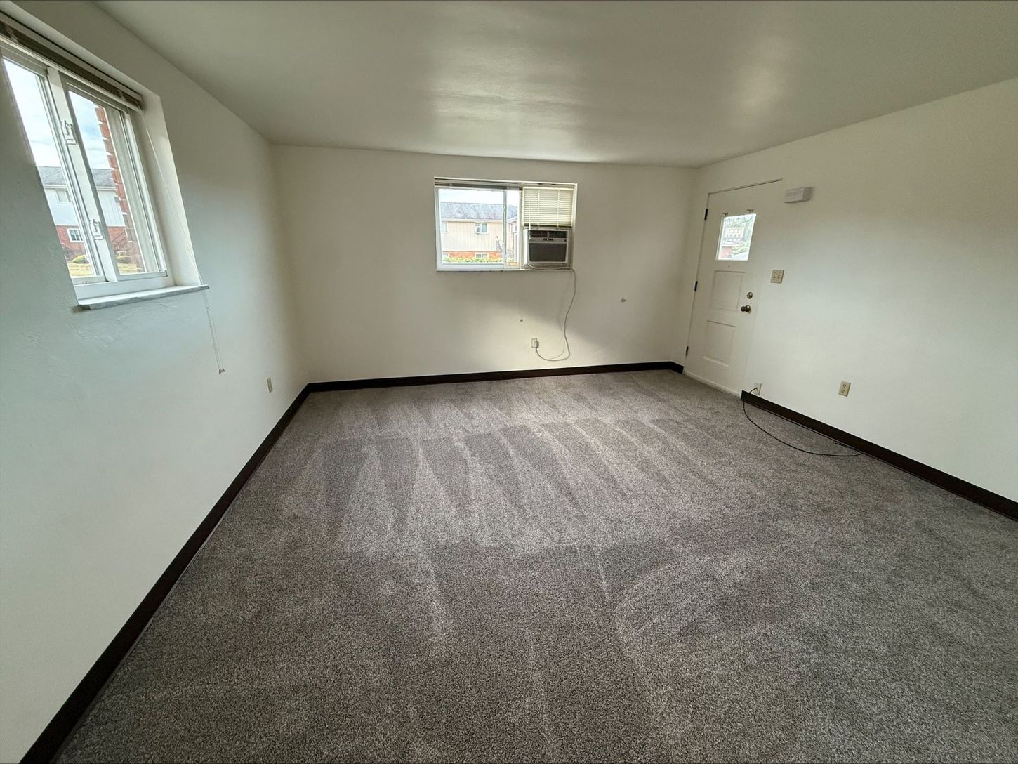 Empty room with gray carpet, two windows, and a door. White walls, natural light.