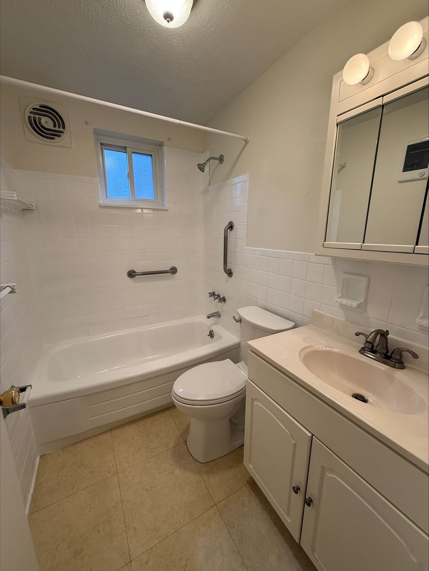 Bathroom with tub, toilet, and vanity. White walls, light-colored floor. Grab bar.