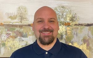 Bald man with goatee smiles, wearing a blue polo. Art piece with gold and browns in background.