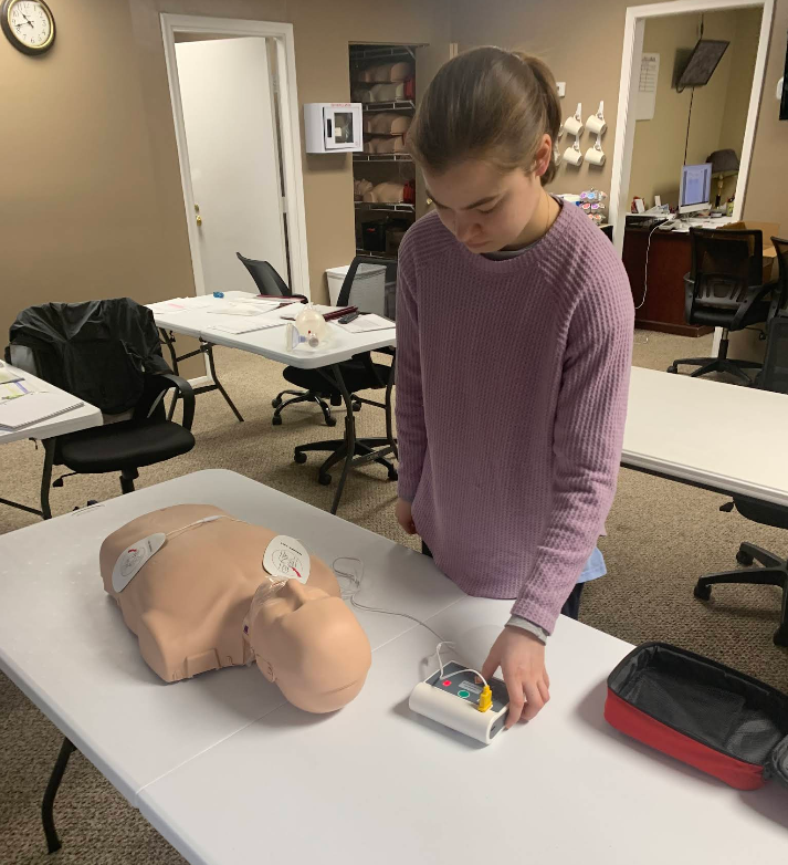 Teenage girl practices CPR with a mannequin and defibrillator on a table.