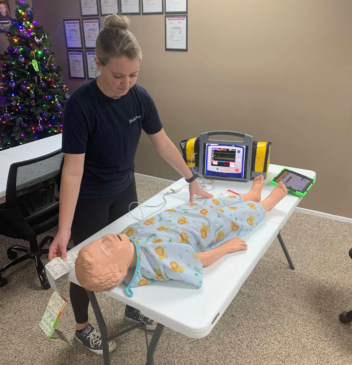 Woman demonstrates CPR on a child mannequin with an AED on a table indoors.