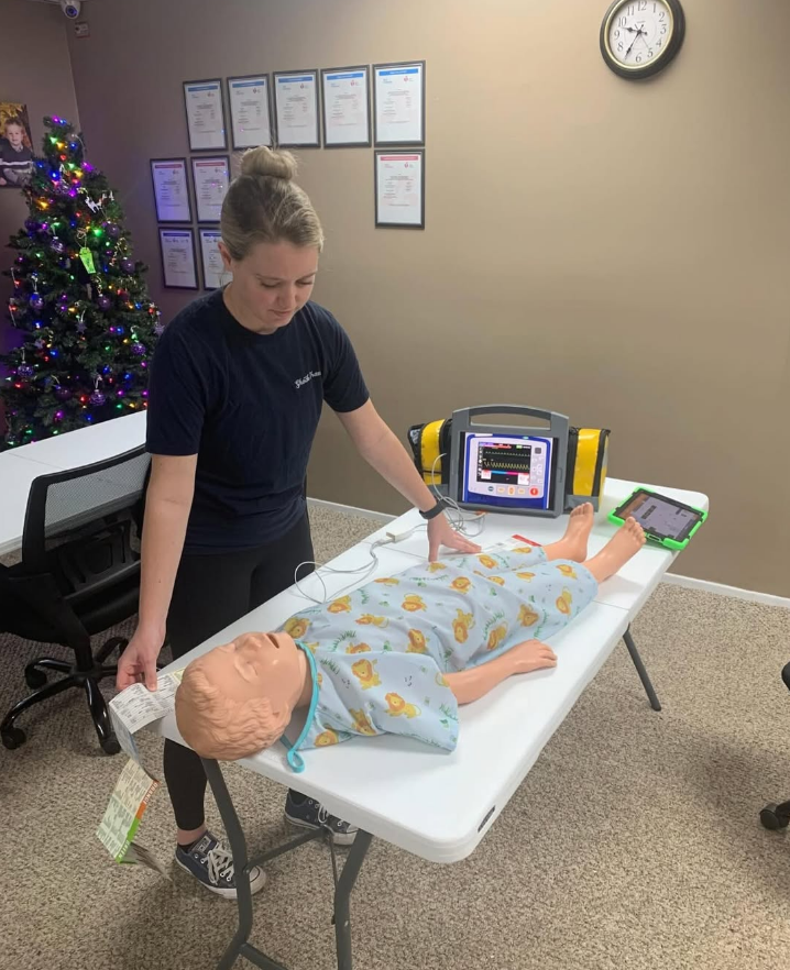 Woman practicing CPR on a baby mannequin, next to a medical monitor on a white table.