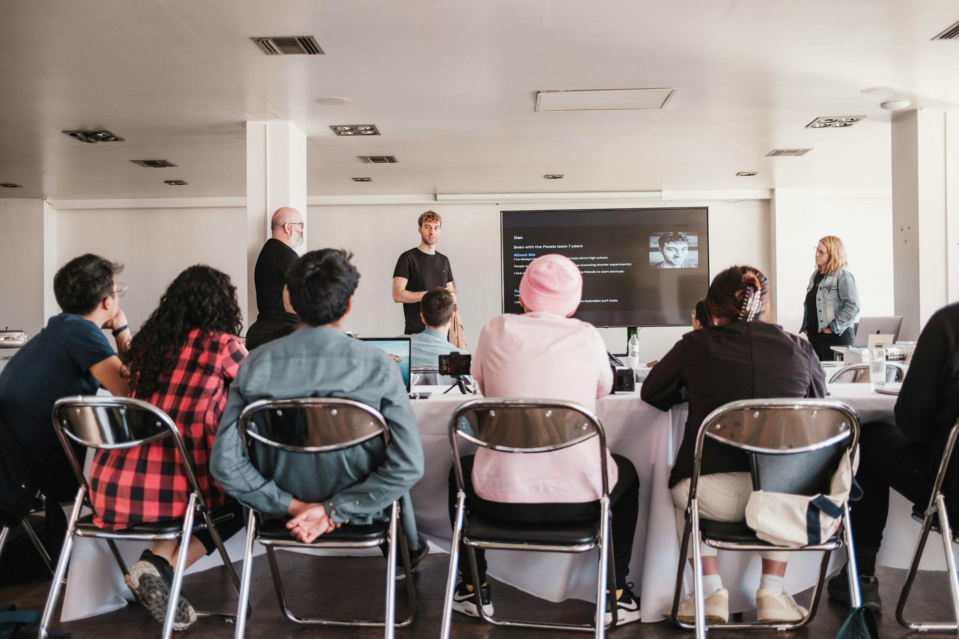 People sit at tables facing a presenter giving a talk in front of a monitor in a bright office space.