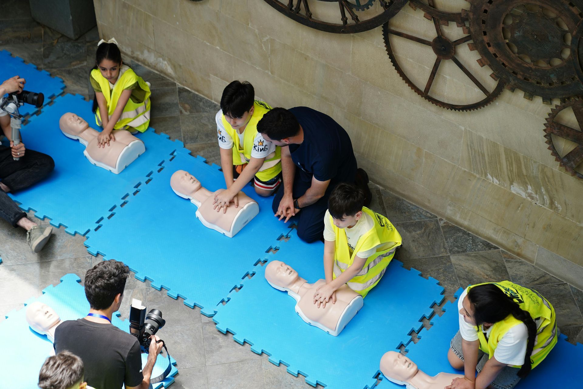 Children in yellow vests practice CPR on mannequins with an instructor; outdoors.