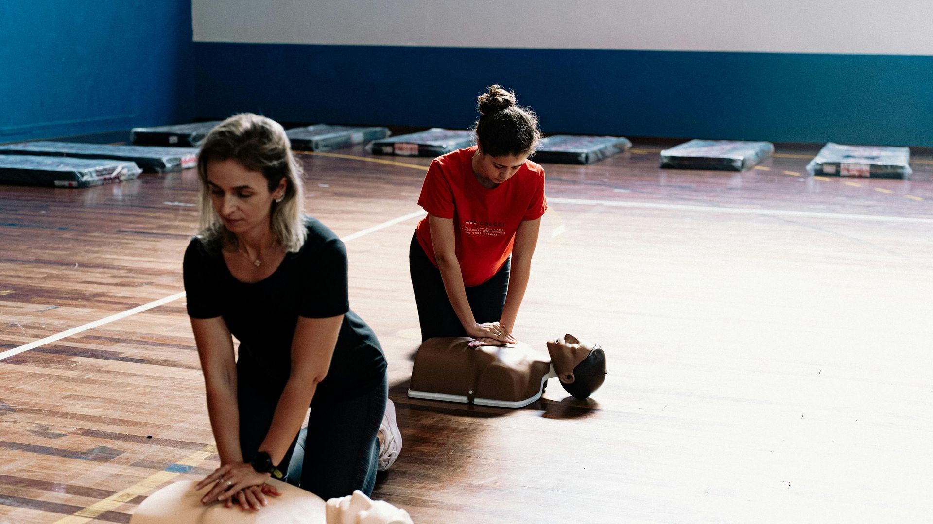 Two people practicing CPR on mannequins in a gymnasium setting.