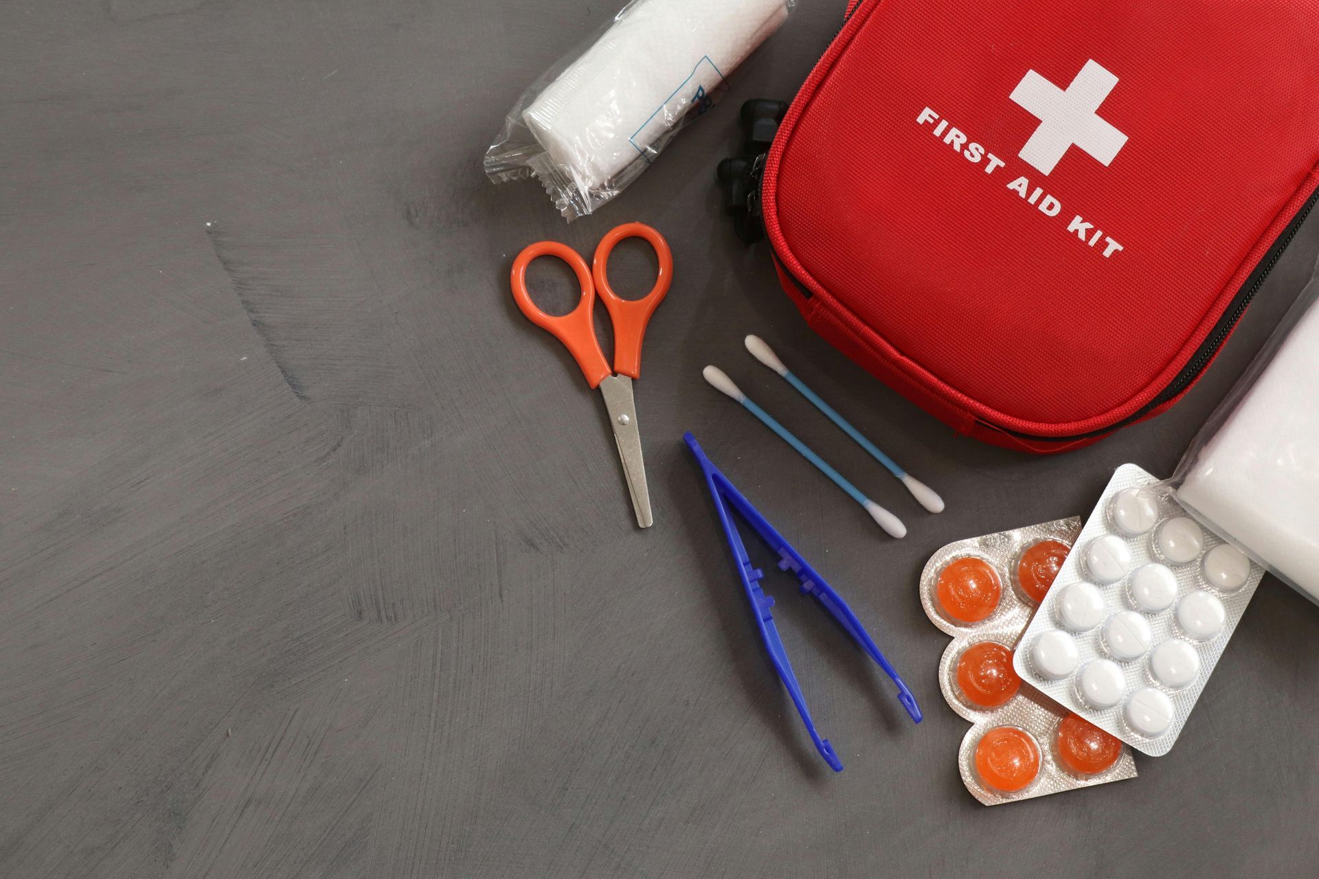 First aid kit, scissors, tweezers, cotton swabs, and blister packs of medicine arranged on a dark grey surface.