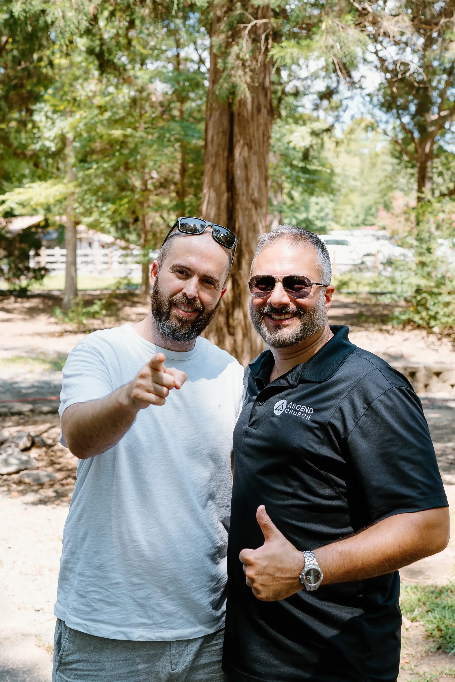Two men are standing next to each other in front of a tree and pointing at the camera.