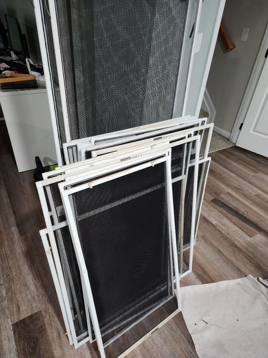Stack of white-framed window screens with black mesh, on a wood-look floor.