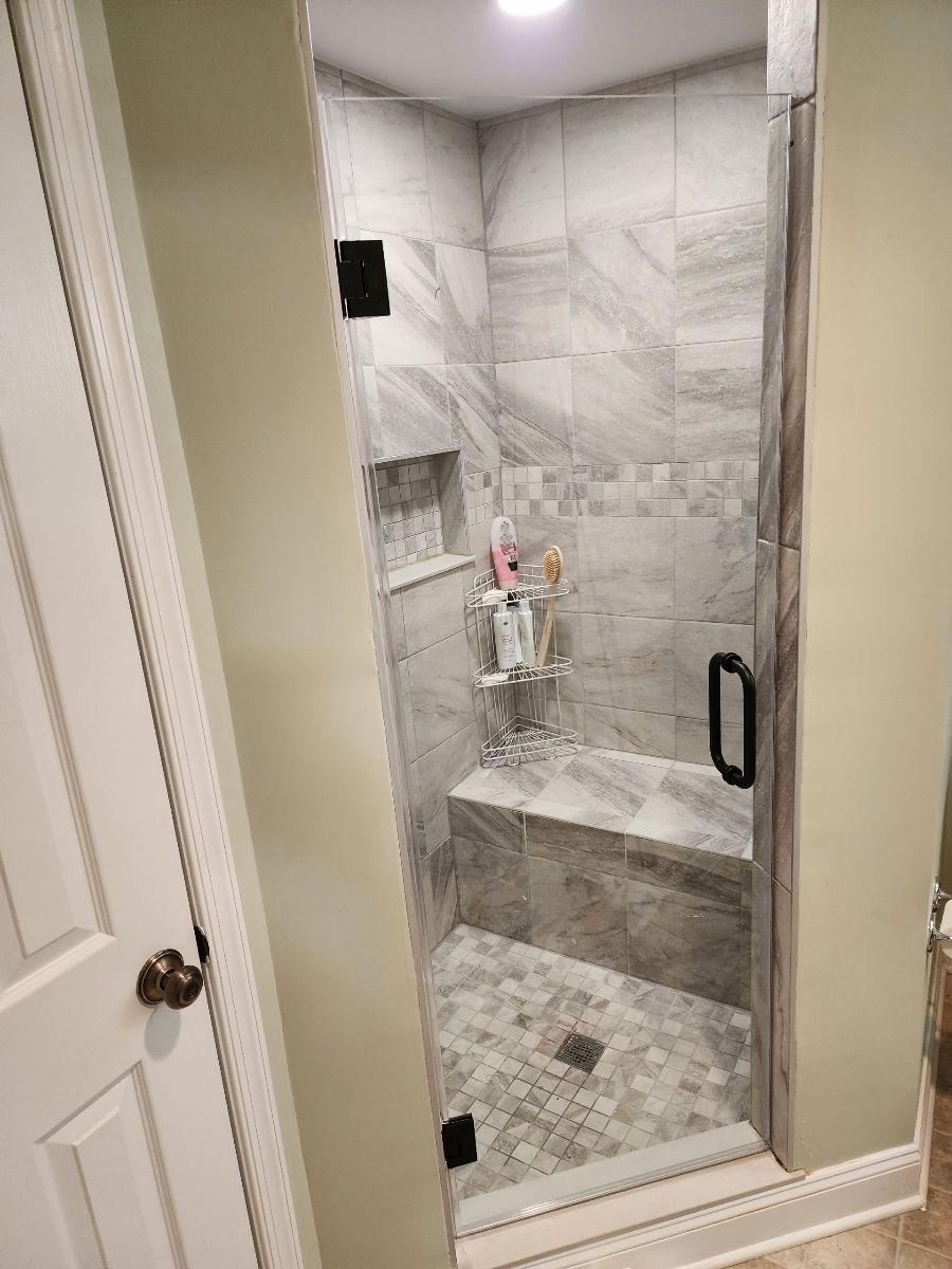 Narrow, tiled shower with a glass door, built-in seat, and shelf for toiletries.