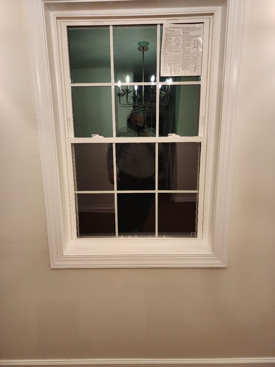 White framed window on a beige wall reflecting a room with a chandelier and a person.