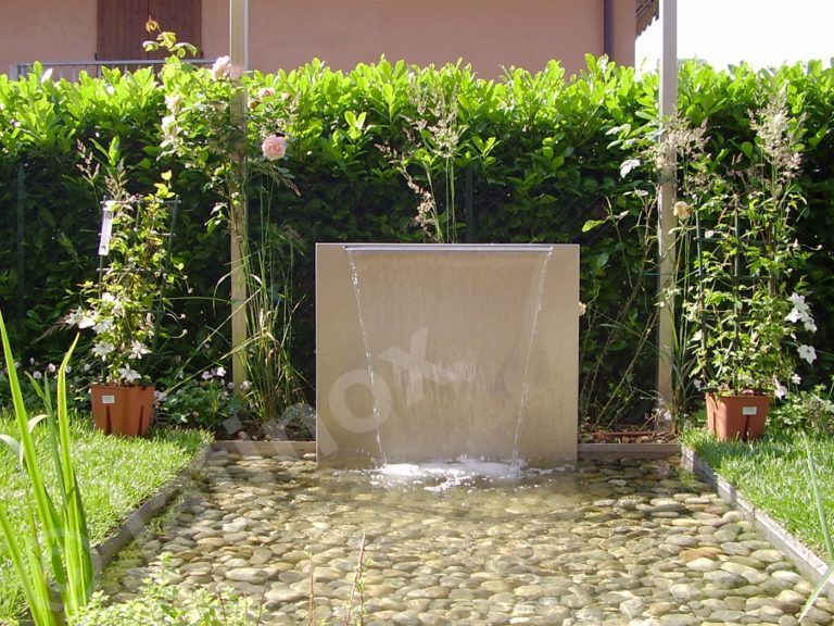 Una moderna fontana da esterno con acqua corrente, circondata da ciottoli, piante e vegetazione.