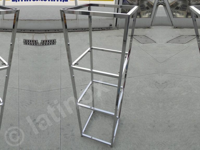 Espositore in metallo, finitura cromata, tre ripiani, su una superficie in cemento grigio.