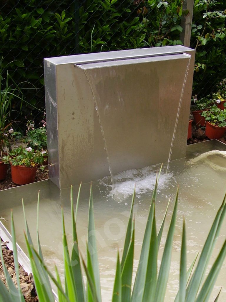 Moderna fontana in metallo in un giardino, acqua che cade a cascata in una piscina poco profonda, verde circostante.