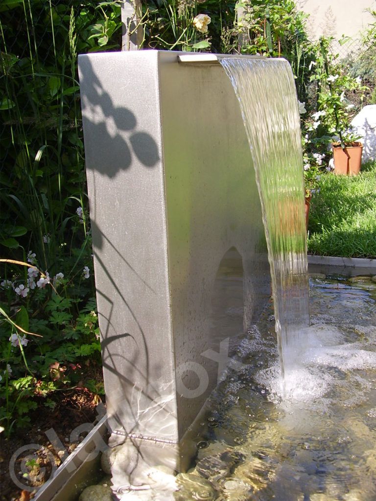 Fontana in acciaio inossidabile con acqua che cade a cascata in una piscina poco profonda, circondata da piante.