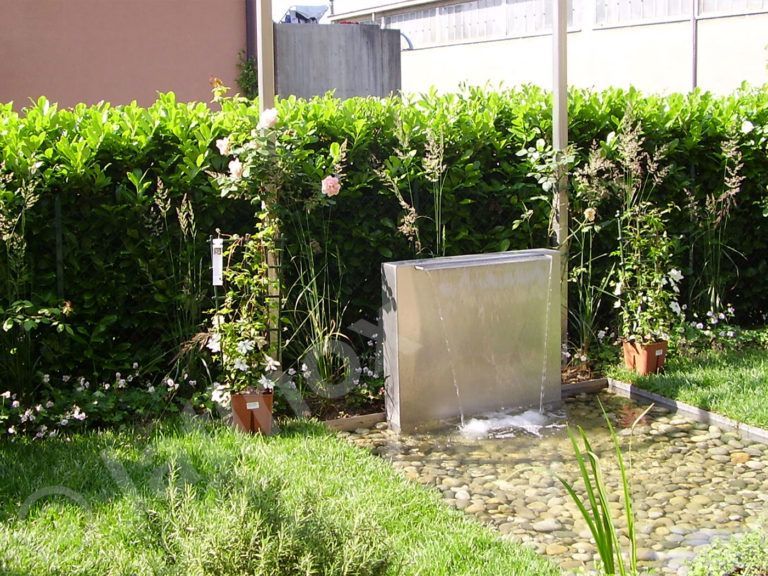 Fontana con pannello quadrato in acciaio inox in un giardino, che scorre in un'area ciottolosa. Sullo sfondo, siepi verdi e rigogliose.
