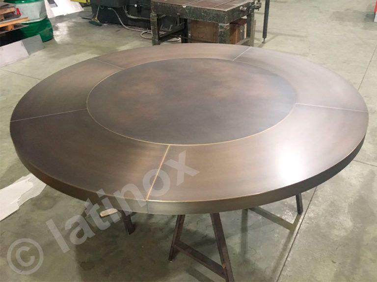 Grande tavolo rotondo in metallo color bronzo con un cerchio centrale rialzato, appoggiato su una struttura metallica in un ambiente da laboratorio.