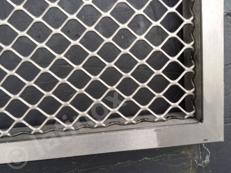 Primo piano di uno schermo in metallo argentato con aperture a forma di diamante, inserito in una cornice metallica.