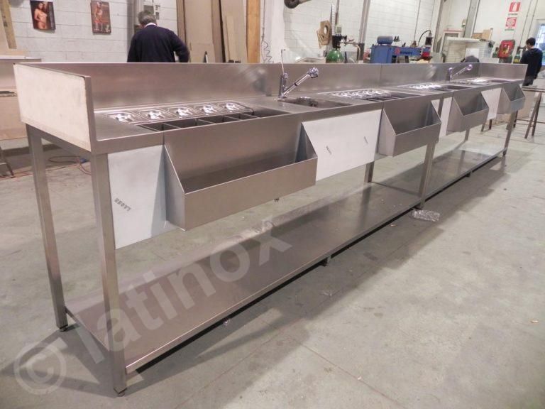 Bancone in acciaio inox con lavelli, contenitori e ripiani, in un ambiente di cucina commerciale.