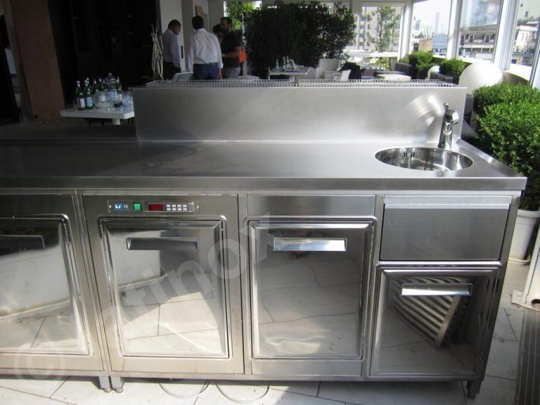 Bar esterno in acciaio inox con lavello, frigoriferi e piano di lavoro, in un ambiente luminoso.