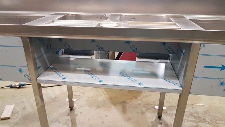 Postazione di lavoro commerciale in acciaio inox con lavelli e ripiano inferiore. Pellicola protettiva blu visibile.