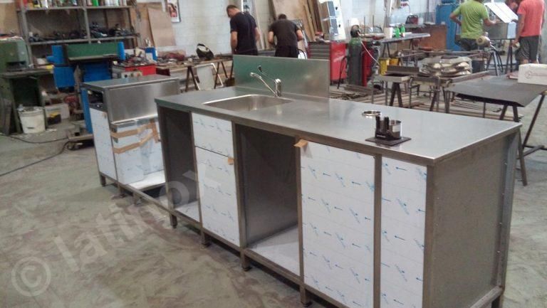 Isola da cucina in acciaio inossidabile in un'officina; operai sullo sfondo.