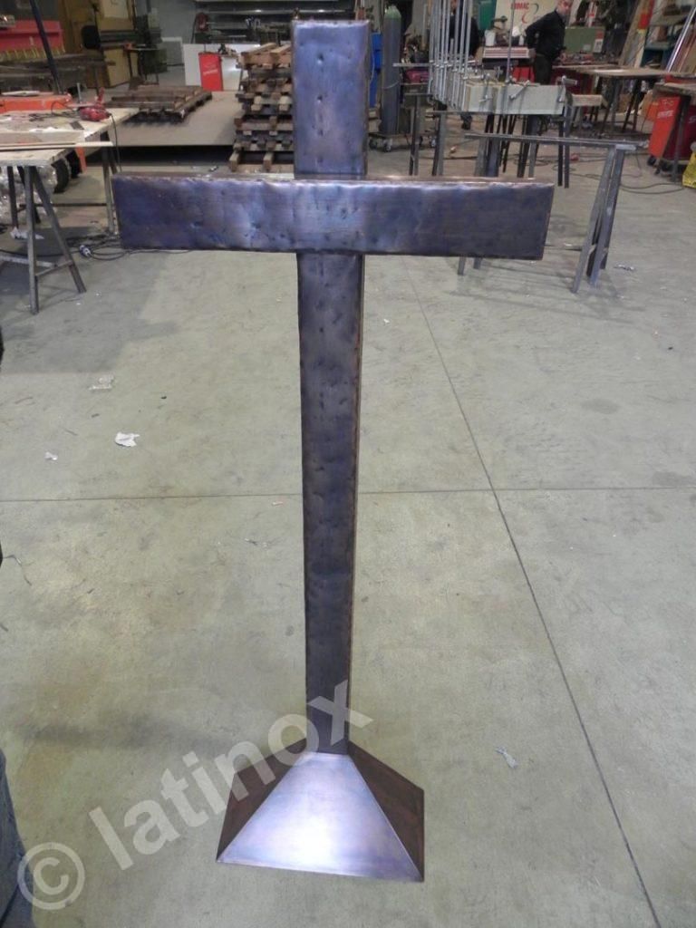 Scultura in metallo a forma di croce con parte superiore rettangolare e base, ambientata in un laboratorio.