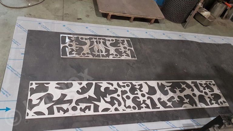 Due pannelli metallici finemente tagliati, uno lungo e uno corto, poggiano su una superficie grigia con del nastro adesivo blu, probabilmente in un'officina.