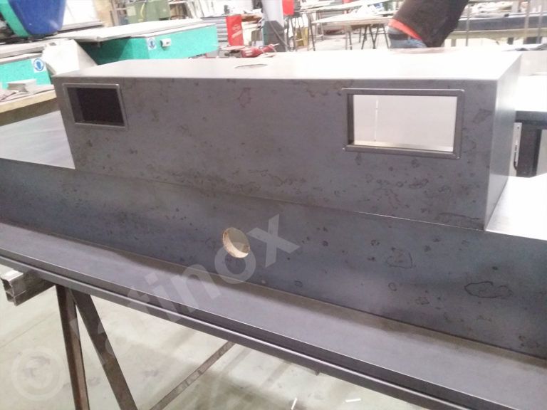 Fabbricazione di metallo: scatola rettangolare grigio scuro con due ritagli rettangolari, appoggiata su un banco da lavoro in metallo.