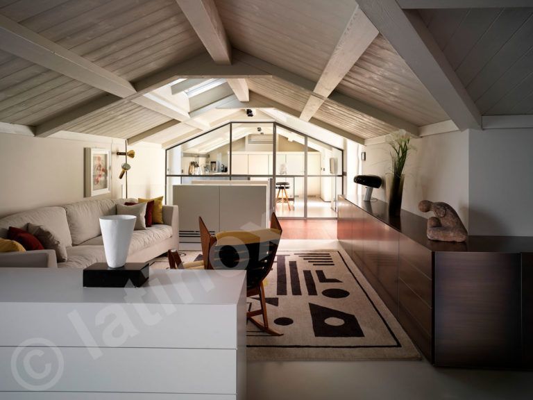 Soggiorno con soffitto spiovente, lucernario, divano beige, cassettiera bianca e tappeto a fantasia.