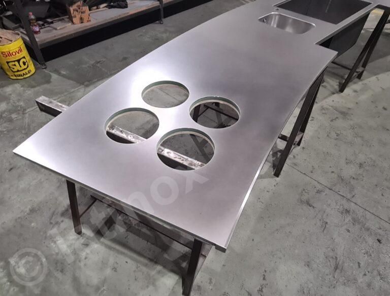 Piano di lavoro in acciaio inox con lavello e quattro intagli circolari, appoggiato su supporti metallici in un'officina.