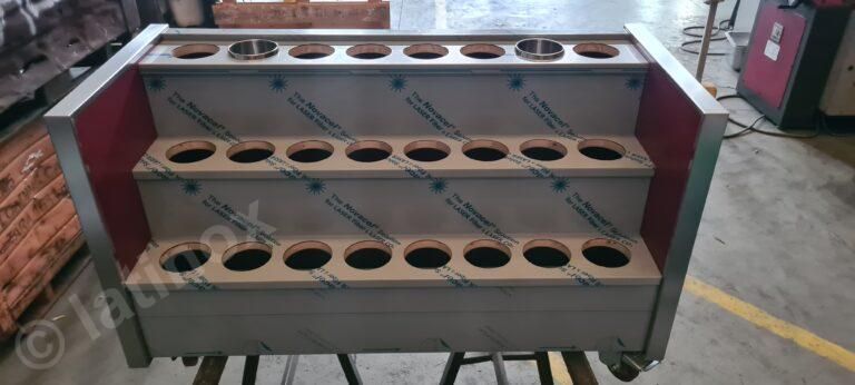 Unità di stoccaggio a più livelli in metallo con fori circolari, probabilmente per bottiglie o contenitori, in un ambiente di fabbrica.