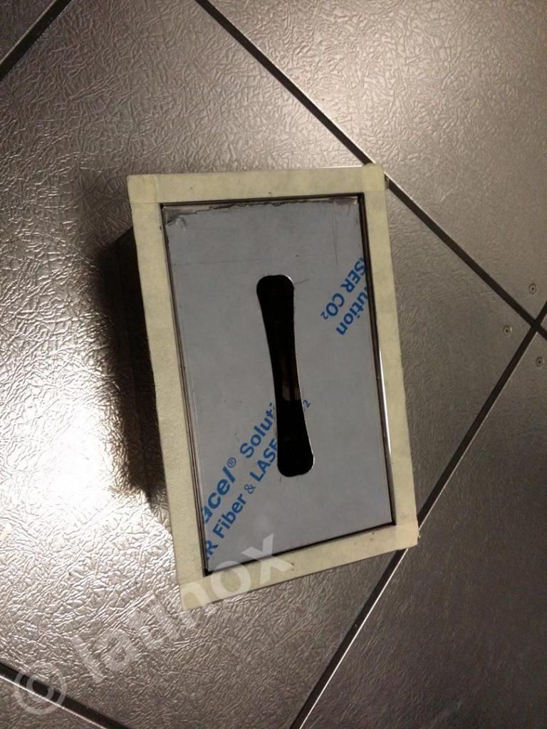 Rettangolo di metallo con ritaglio, fissato con nastro adesivo a una superficie più scura.