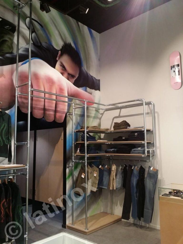 Interno di un negozio di abbigliamento con scaffali cromati che espongono camicie e pantaloni. Un grande disegno raffigura un uomo che indica.