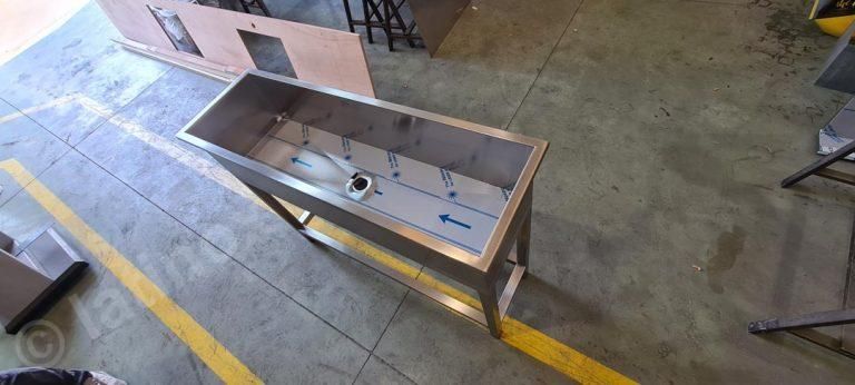 Lavello industriale in acciaio inossidabile su una struttura metallica, in un'officina, vicino a una linea gialla sul pavimento.