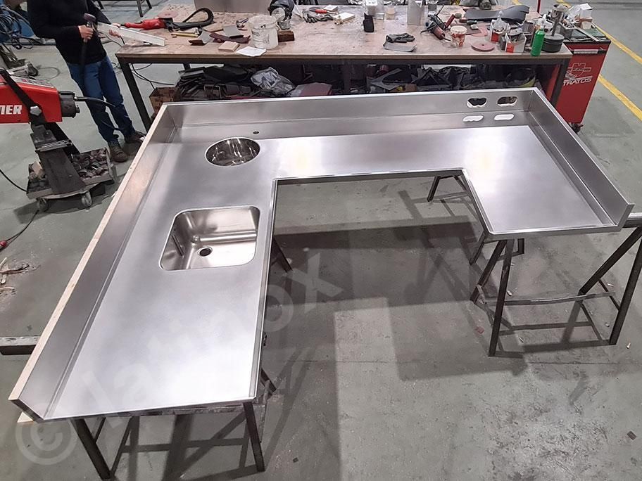 Piano di lavoro a U in acciaio inox con fori per il lavello, posizionato su supporti metallici in un'officina.