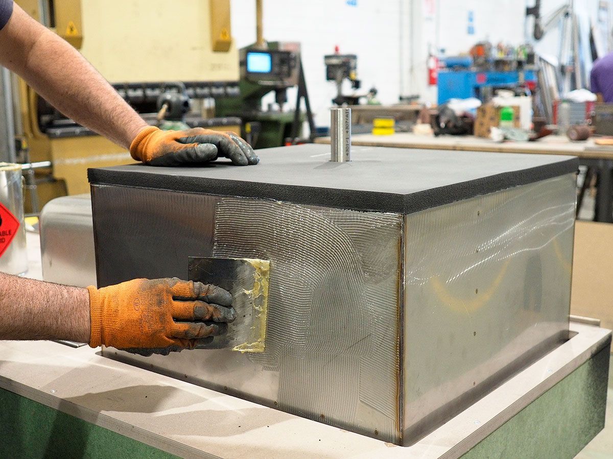 Due operai applicano l'adesivo con una spugna su una scatola di metallo con il coperchio di schiuma grigia, in un'officina.