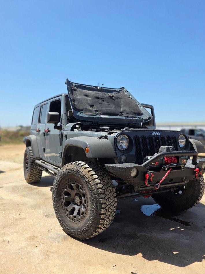 A jeep with its hood up is parked in the dirt.