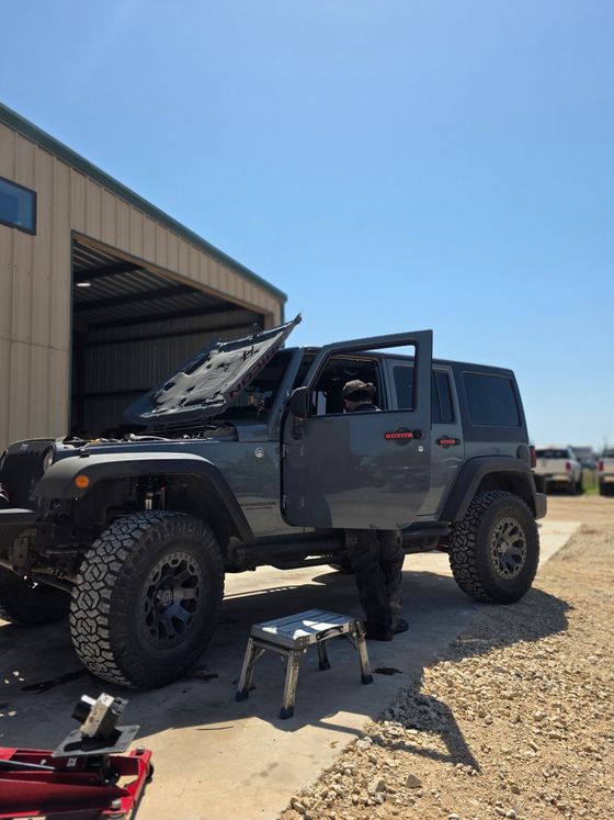 A man is working on a jeep with its hood up in front of a garage.