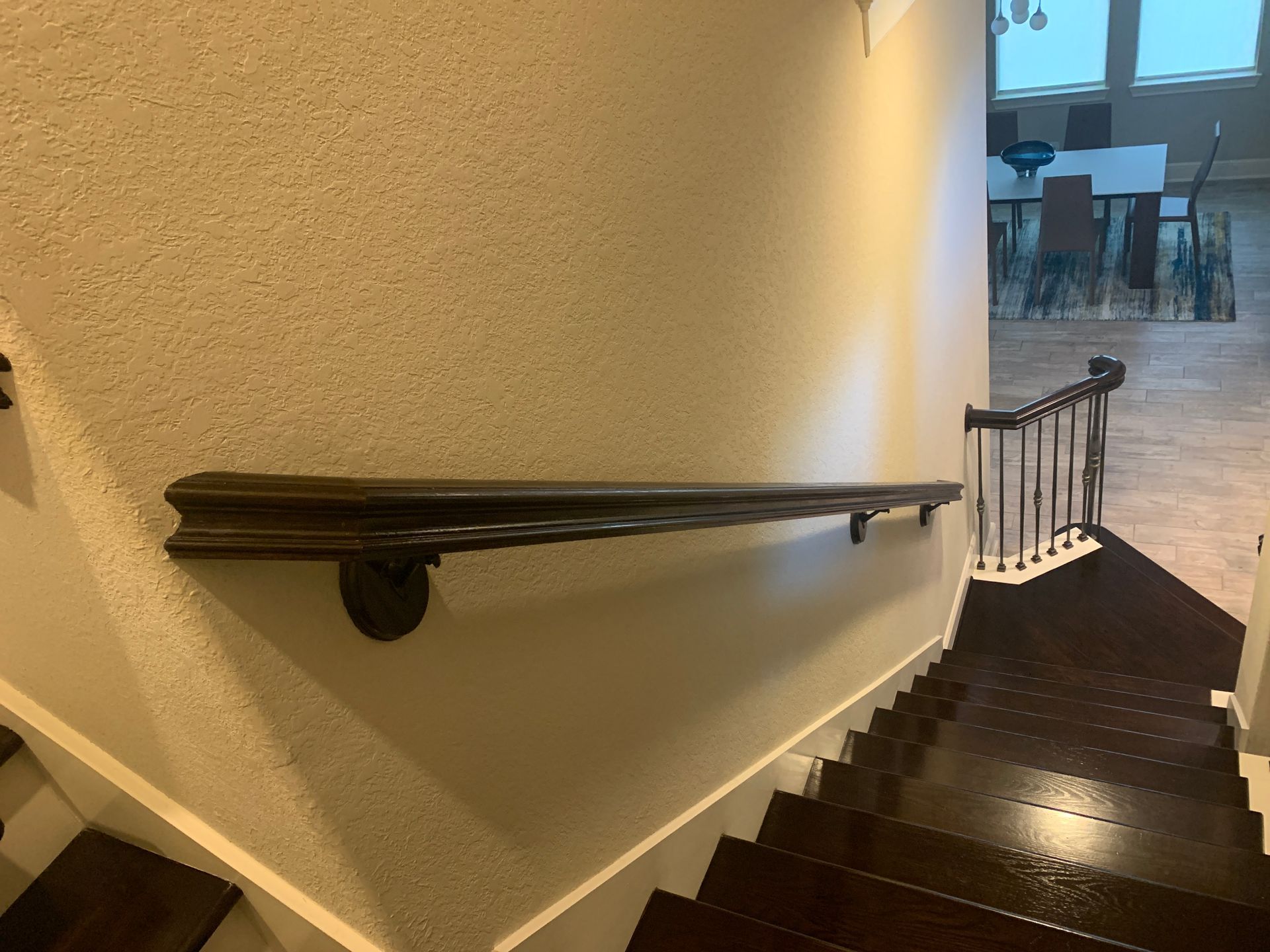 A dark wood handrail mounted on a beige wall alongside a carpeted staircase leading down to a dining area.
