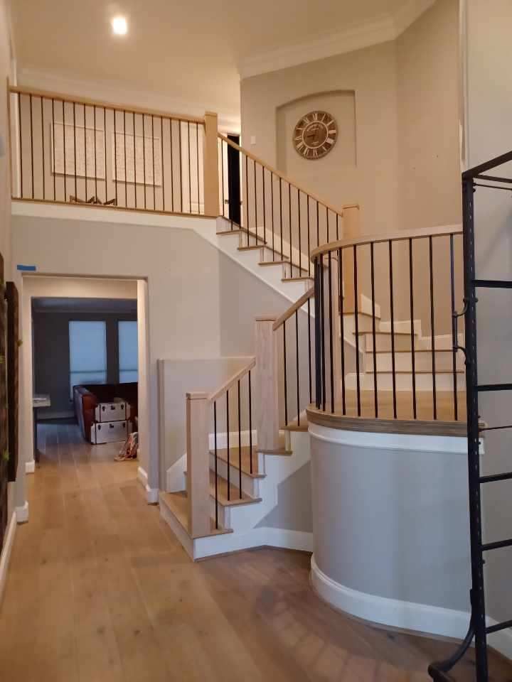 A grand foyer features a curved staircase with dark metal railings and light wood flooring under soft, neutral wall tones.