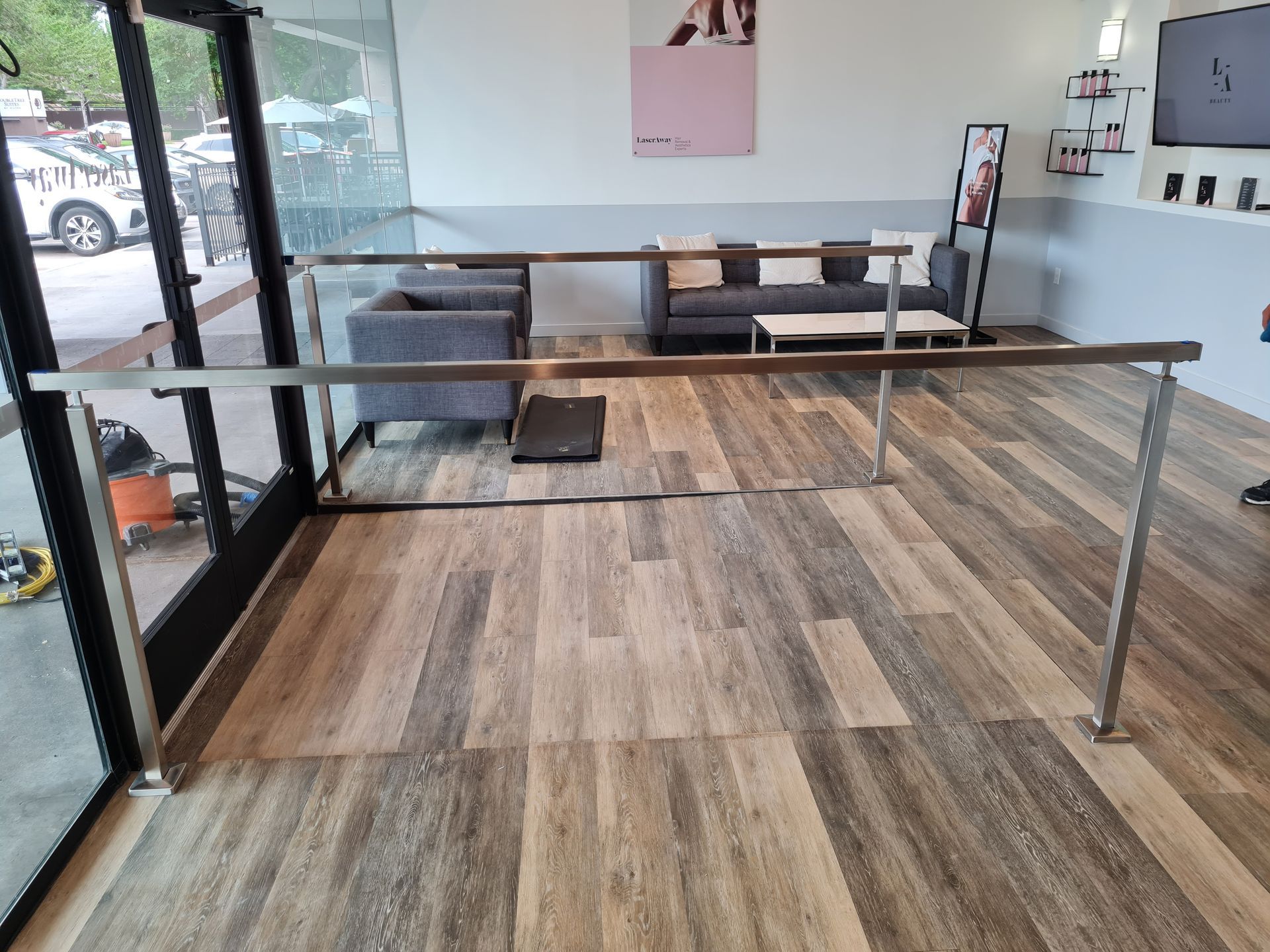 Interior view of a waiting area with wood-look flooring, a gray sofa, a small coffee table, and a metal safety handrail.