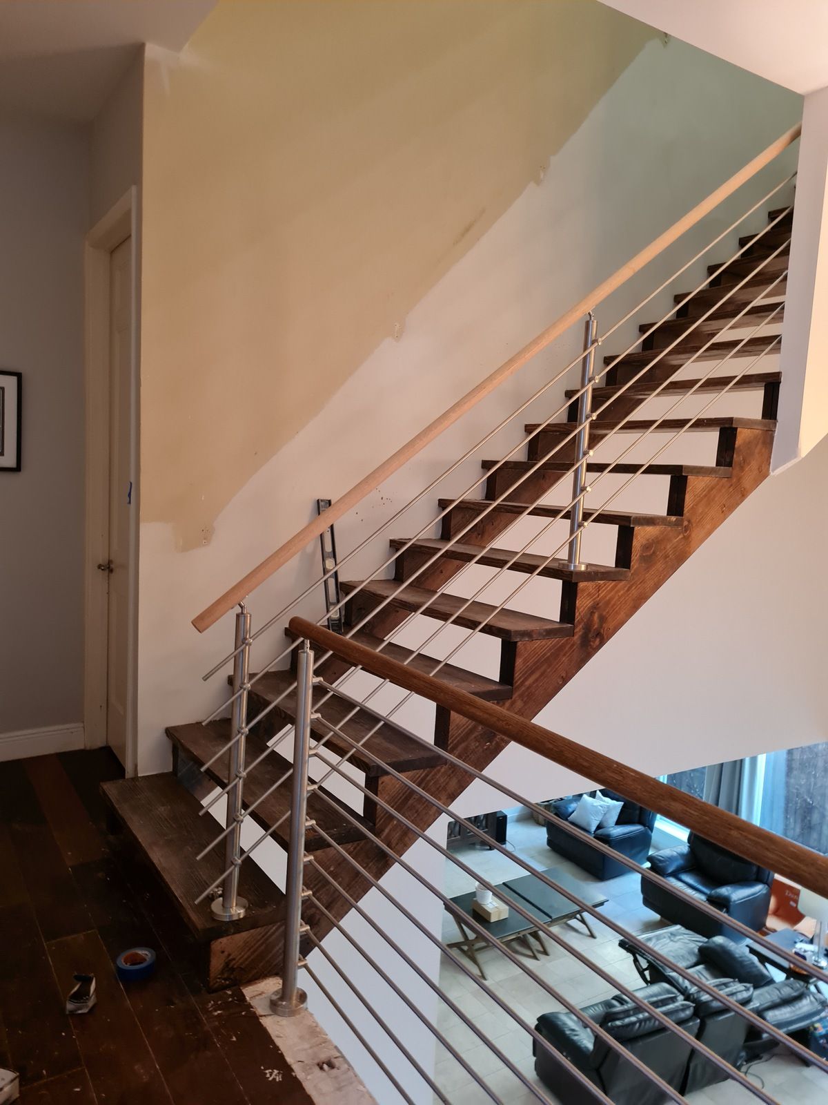 A wooden staircase with metal cable railing ascends next to a half-painted wall, overlooking a living area below.