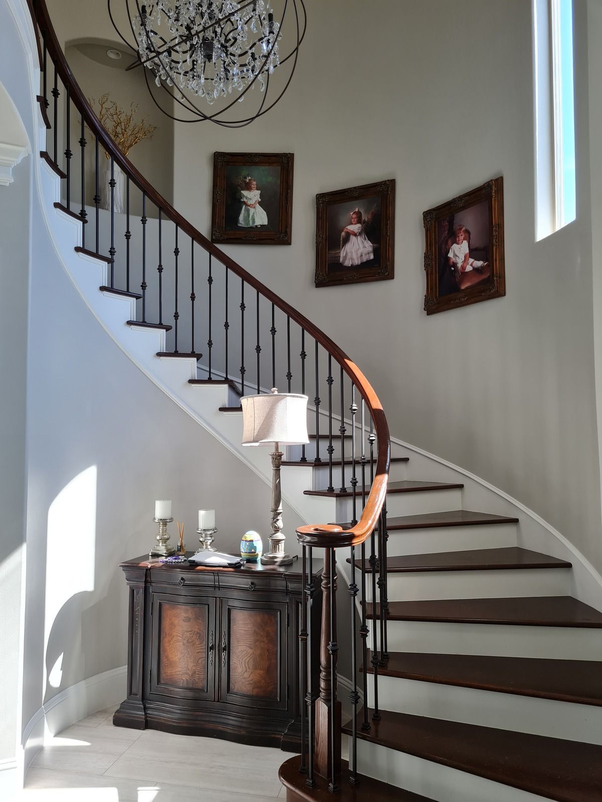 A curved staircase with wooden steps and metal railings, leading to a wall with three portraits and a side cabinet below.