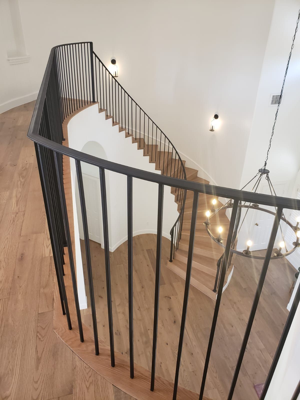 A high-angle view of a curved staircase with black metal railings, wooden steps, and a chandelier in a white hallway.