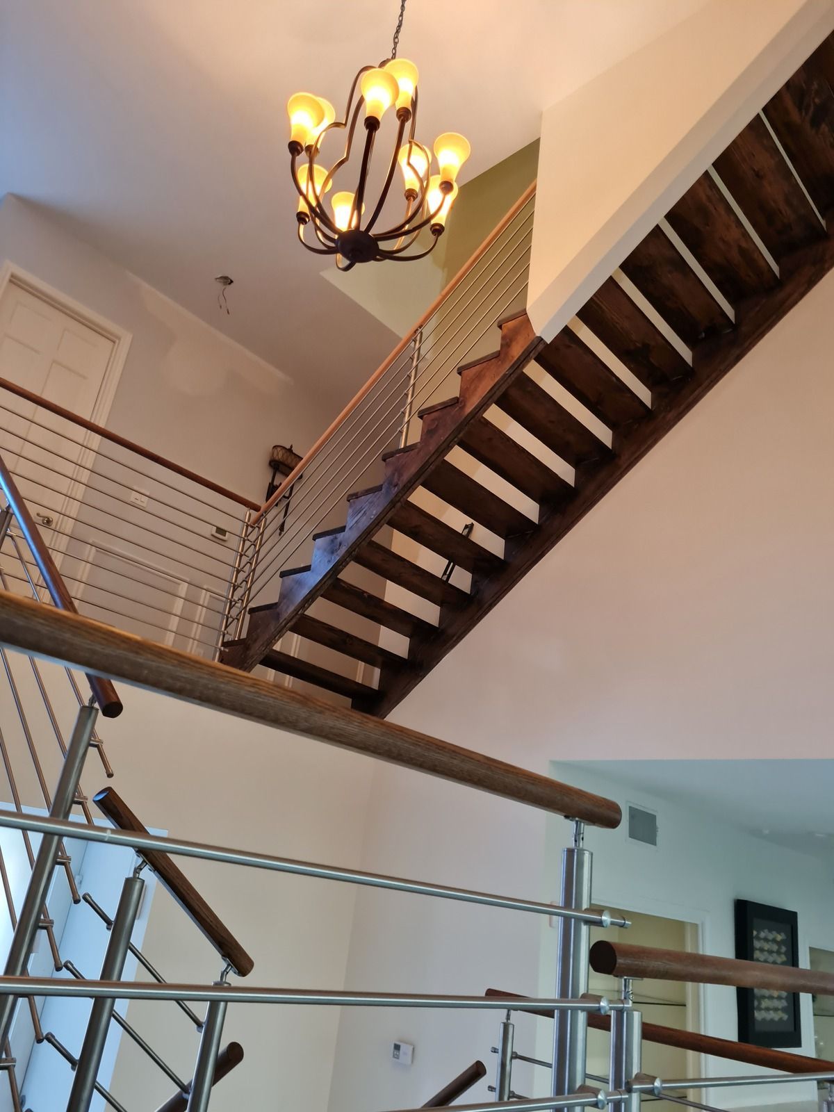 A modern wooden staircase with cable railings ascends through an open, high-ceilinged room with a decorative chandelier.